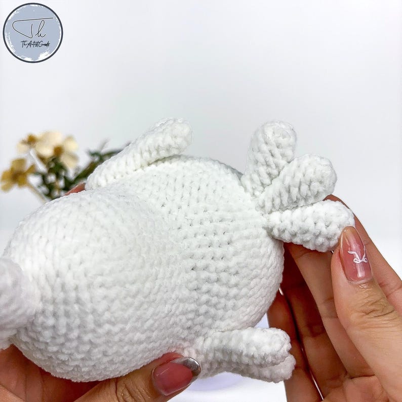 May include: White crocheted stuffed animal with a rounded body and a tail with three points. The animal is being held by a hand.