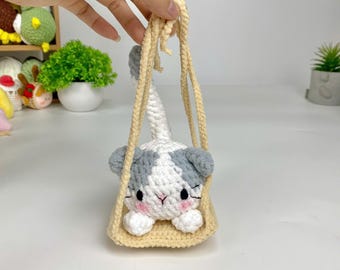 May include: A crocheted cat toy hanging from a cream-colored crocheted swing. The cat is white with gray patches and has pink cheeks.