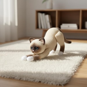 以下が含まれることがあります： 白いラグの上でストレッチポーズをとる、かぎ針編みのシャム猫。猫はクリーム色の体で、顔、耳、足に茶色の模様があります。青い目と白い足を持っています。猫は遊び心のある姿勢です。