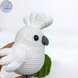May include: White crocheted cockatoo plush toy with a gray beak and black eyes. The toy is sitting on a green crocheted surface.