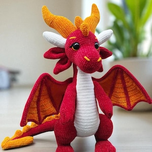 May include: A handmade crochet dragon in shades of red, orange, and white. The dragon has large wings, horns, and a long tail. The dragon is standing upright, with its wings spread slightly. The dragon is a cute and whimsical toy.