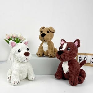May include: Three crocheted dog figurines in white, tan, and brown. The white dog has pink ear accents, the tan dog has a black nose, and the brown dog has white markings. All three are sitting with their paws forward.