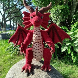 May include: A handmade red crochet dragon with brown horns, wings, and claws. The dragon has a cream-colored belly and is posed on a large gray rock. The dragon is a whimsical, handcrafted item.