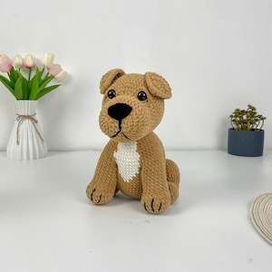 May include: A handmade, light brown crochet dog with black eyes and a black nose. The dog has a white patch on its chest and is sitting upright. The dog is a stuffed toy, likely for a child.