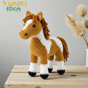 May include: A crocheted horse toy, approximately 4 inches (10 cm) tall, in shades of brown, white, and black. The horse has a white blaze on its face and a white patch on its body. The mane and tail are brown.