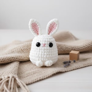 May include: A white crochet bunny with pink inner ears and black button eyes. The handmade stuffed animal sits on a beige scarf. A small wooden cube and dried lavender sprigs are next to the bunny.