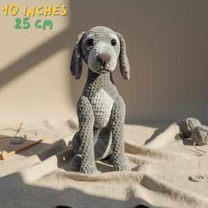 Può includere: Un cane all'uncinetto grigio fatto a mano, alto 25 cm, con petto bianco e naso marrone. Il cane è seduto su un tessuto beige. Il testo "10 inches 25 cm" è in alto a sinistra.