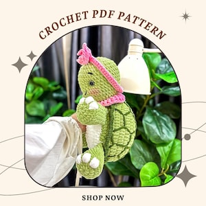 Peut inclure: Une tortue crochetée avec une carapace verte, des pieds blancs et un bandeau et une écharpe roses. La tortue tient un objet blanc. L'image comprend le texte "CROCHET PDF PATTERN" et "SHOP NOW".