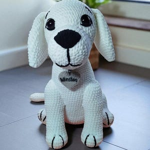 Puede incluir: Un perro blanco de ganchillo hecho a mano con detalles negros en la nariz, los ojos y las patas. El perro tiene orejas caídas y una etiqueta en forma de corazón que dice "Winston". El peluche está sentado en un suelo de madera.