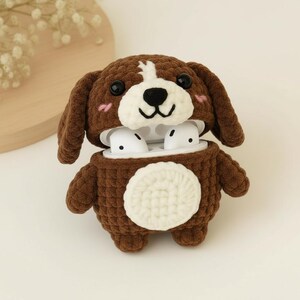 May include: A brown and white crocheted dog-shaped case for wireless earbuds. The case has a white belly, black eyes, and pink cheeks. The earbuds are white. The case is designed to protect and carry the earbuds.
