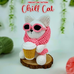 May include: White crocheted cat wearing pink sunglasses and a pink scarf, holding a crocheted cup of coffee. The cat is sitting on a wooden base. The text "Chill Cat" is written in red above the cat.
