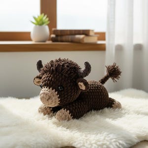 May include: A handmade brown crochet bull plush toy with a light brown face and hooves, lying on a white fluffy rug. The bull has small horns and a fluffy tail. The background includes a window, books, and a small potted plant.