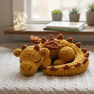 May include: A crocheted dragon plush toy in shades of yellow and brown, lying on a white textured surface. The dragon has closed eyes and a curled tail, with brown accents on its horns and spine. The toy is soft and cuddly.