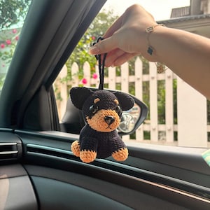 Puede incluir: Un juguete de perro de crochet negro y marrón colgando del espejo retrovisor de un coche. El juguete tiene un cuerpo negro y una cara y patas marrones.