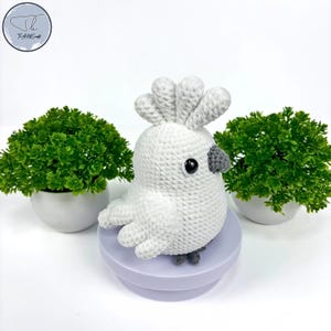 May include: White crocheted cockatoo bird with gray beak and black eyes. The bird is sitting on a purple base.