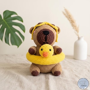 May include: A brown crocheted bear toy wearing a yellow swimming ring with a small yellow duck head attached to the front. The bear has a dark brown snout, black eyes, and a yellow headband. The texture is soft and plush with visible yarn loops.
