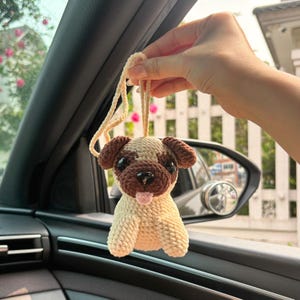 Puede incluir: Un perro pug de crochet colgando del espejo retrovisor de un coche. El pug es beige con ojos marrones y una lengua rosa que sobresale.