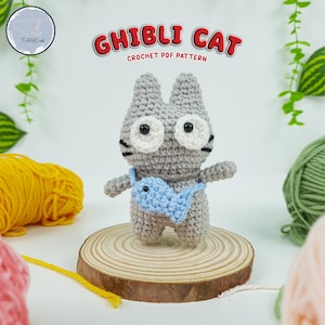 May include: A gray crocheted cat with white eyes and a blue fish in its mouth. The cat is sitting on a wooden base. The text "GHIBLI CAT CROCHET PDF PATTERN" is above the cat.
