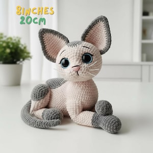 Puede incluir: Muñeca de gato de ganchillo, de unos 20 cm de largo, en posición sentada. El gato es de color rosa claro con detalles grises en las orejas, la cabeza y las patas. Tiene grandes ojos azules y bigotes blancos. El texto "8 inches 20cm" es visible.