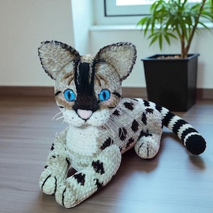Peut inclure: Une peluche faite à la main représentant un chat aux yeux bleus, au pelage beige et aux taches noires. Le chat a de grandes oreilles et une queue rayée noire et blanche. Le jouet est couché, avec des détails soignés.