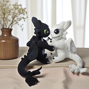 Op de afbeelding: Twee gehaakte drakenfiguren, één zwart en één wit, staan samen. De zwarte draak heeft groene ogen, terwijl de witte draak blauwe ogen heeft. Beide hebben vleugels en zijn gemaakt van zacht garen. De draken zijn ongeveer 15 cm hoog.