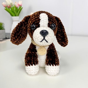 May include: A handmade crochet dog toy in brown, white, and beige. The plush dog has large black eyes, floppy ears, and a small beige collar with a round tag. The dog is sitting upright, showcasing its detailed craftsmanship.