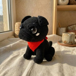 May include: A handmade black crochet pug dog with large, expressive eyes and a bright red bandana. The plush toy is sitting on a neutral-colored surface. The pug has a cute, friendly expression.