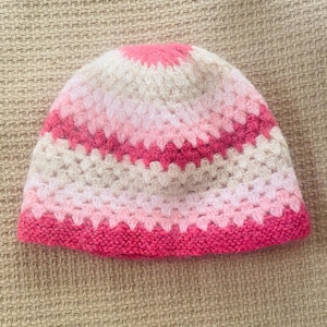 May include: A crochet beanie featuring a striped pattern in shades of hot pink, light pink, and cream. The hat is constructed with a textured granny square stitch pattern throughout the body and finished with a solid hot pink ribbed knit edge.