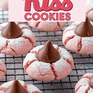 K&ouml;nnte beinhalten: Rosa und wei&szlig;e Kekse mit Schokoladenk&uuml;ssen darauf, auf einem Gitterrost angeordnet. Die Kekse sind mit Puderzucker best&auml;ubt und der Text "Strawberry Kiss Cookies" ist auf rosa Hintergrund dargestellt.