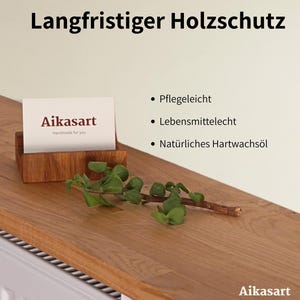 Ein hölzerner Visitenkartenhalter mit einer Karte mit der Aufschrift "Aikasart Handmade for you" steht auf einer Holzoberfläche. Der Text "Langfristiger Holzschutz" steht oben. Ein Zweig mit Grün liegt auf dem Holz.