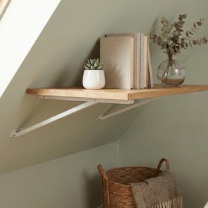 Op de afbeelding: Een licht houten zwevend plankje met witte metalen beugels aan de muur. Op het plankje staan boeken, een kleine plant in een pot en een glazen vaas met gedroogde takken. Een rieten mand staat eronder.