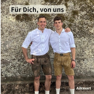 May include: Two individuals wearing white button-down shirts and leather lederhosen shorts. One pair of shorts is dark brown, and the other is light tan. Both feature intricate embroidery. The text "F&uuml;r Dich, von uns" appears at the top, and "Aikasart" at the bottom.
