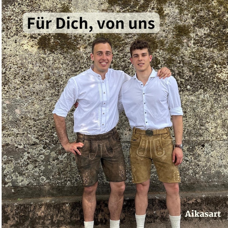 May include: Two individuals wearing white button-down shirts and leather lederhosen shorts. One pair of shorts is dark brown, and the other is light tan. Both feature intricate embroidery. The text "F&uuml;r Dich, von uns" appears at the top, and "Aikasart" at the bottom.