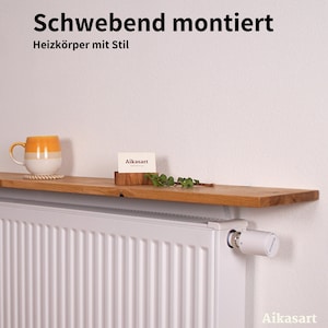 Massives Eichenholzbrett als Heizkörperablage, schwebend auf einem weißen Heizkörper montiert. Oben stehen eine Pflanze, eine Kerze und ein Buch. Die natürliche Maserung und das geölte Finish machen die Ablage zu einem stilvollen Deko-Element im Wohnraum.