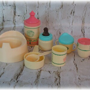 May include: A set of vintage plastic baby doll accessories in cream, pink, and blue. The set includes a potty, a bottle, a cup, a bowl, and a spoon. The items are decorated with a green and white floral pattern.