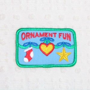 May include: A green, blue, and red embroidered patch with the text "ORNAMENT FUN". The patch features a red stocking, a yellow star, and a red heart with a yellow center.