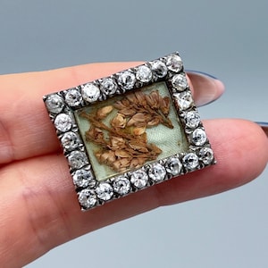 Pode incluir: Broche quadrado e antigo com uma moldura prateada cravejada de pedras preciosas redondas e transparentes. O centro apresenta um fundo verde claro com matéria vegetal seca. Acessório vintage.