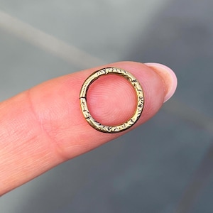 May include: A small, gold-colored hoop earring with an etched floral pattern. The earring is circular and appears to be hinged for easy opening and closing. The jewelry is held between two fingers.