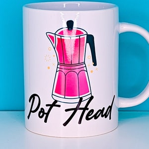 Könnte beinhalten: Weiße Keramik-Tasse mit einer rosa Abbildung einer Espressokanne. Auf der Tasse steht in schwarzer Schrift "Pot Head". Der Hintergrund ist einfarbig blau.