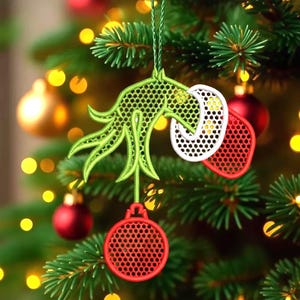 May include: A green, white, and red Christmas ornament with a whimsical design, hanging on a Christmas tree. The ornament features a green hand-like shape, a white circle, and a red mitten-shaped element, all with a perforated pattern. A red ball-shaped ornament hangs below.
