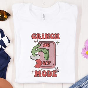 May include: White t-shirt with a retro design featuring the text "GRINCH ON OFF MODE". The graphic shows a green hand in a red sleeve turning a switch. The design is in red, pink, and green.