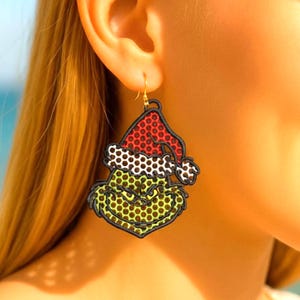 May include: A pair of novelty earrings featuring a green character with a red and white Santa hat. The earrings have a black outline and a gold-colored hook. The character's face is green with a yellow-green background.