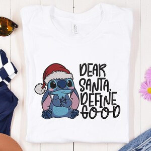 May include: White t-shirt featuring a blue cartoon character wearing a Santa hat. The text "DEAR SANTA. DEFINE GOOD" is printed in black. Sunglasses and other items are visible in the background.