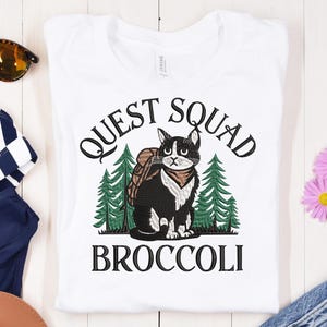 Op de afbeelding: Wit T-shirt met een zwart-witte kat die een rugzak draagt en omringd is door groene bomen. De tekst "QUEST SQUAD BROCCOLI" is op het T-shirt gedrukt.