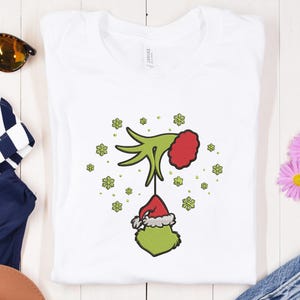 May include: White t-shirt featuring a green hand holding a red and white Santa hat with a green face. The design is surrounded by green snowflakes. Sunglasses and a blue and white striped item are visible in the background.