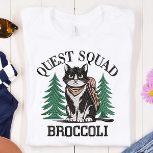 Op de afbeelding: Wit T-shirt met een zwart-witte kat die een rugzak en een bandana draagt. De kat staat voor twee groene dennenbomen. De tekst "QUEST SQUAD BROCCOLI" is op het T-shirt gedrukt.
