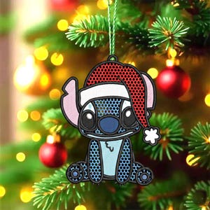 Peut inclure: Ornement de Noël représentant le personnage Stitch de Lilo & Stitch, portant un bonnet de Père Noël. L'ornement est bleu, rose et noir avec un bonnet de Père Noël rouge et blanc. Il est suspendu à une ficelle verte sur un fond flou de sapin de Noël.