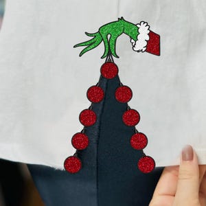 Pode incluir: Camiseta branca com um desenho de árvore de Natal. A árvore é feita de enfeites vermelhos brilhantes e uma base azul escura. Uma mão verde brilhante com um punho vermelho e branco segura a árvore.