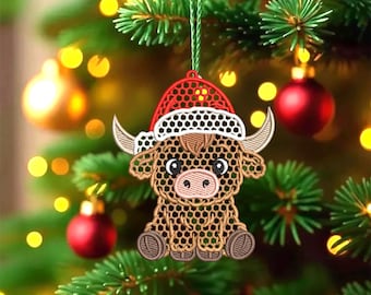 May include: Embroidered Christmas ornament featuring a Highland cow wearing a Santa hat. The ornament is brown, tan, and white with a red and white hat. It is hanging from a green string against a blurred background of a Christmas tree with red ornaments and yellow lights.
