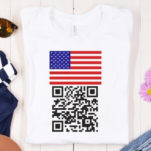 May include: White t-shirt with a design of the American flag and a QR code. The flag has red and white stripes and a blue section with white stars. The QR code is black and white.
